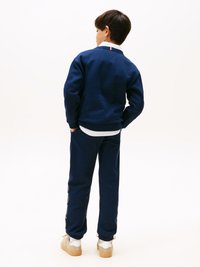 Felpa blu navy e pantaloni sportivi coordinati con polsini elasticizzati, t-shirt bianca sotto e scarpe beige; colletto della camicia visibile all'scollatura.