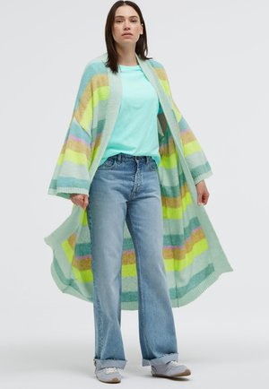 Frau trägt hellblaues T-Shirt, Jeans mit weitem Bein, graue Turnschuhe und langen, mehrfarbigen gestreiften Cardigan mit offenem Vorderteil.