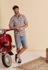 Korte mouwen, roze bloemen knoopshirt, denim shorts, marineblauwe sneakers; vintage rode scooter in de buurt, tan achtergrond, gestreepte vloerkleed eronder.