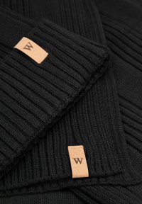 Černá pletená šála s žebrovanou texturou, zdobená malou světle hnědou koženou cedulkou s vyraženým písmenem "W". Materiál je měkký a pružný.