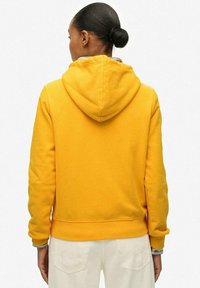 Felpa gialla con cappuccio e texture morbida, dotata di cappuccio regolabile e polsini a coste. Il capo è abbinato a pantaloni di colore chiaro.