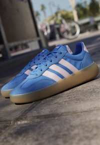 Blue Adidas sneakers with pink stripes and gum soles placed on a concrete surface outdoors with blurred background.