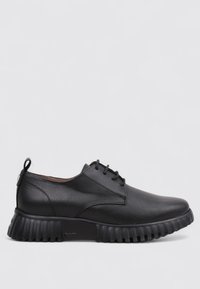 Zapato negro de cuero con cordones, con parte superior texturizada y suela de goma gruesa y estriada, contra un fondo gris claro.