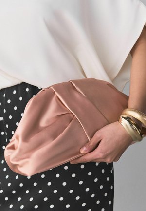 Osoba trzyma różową satynową kopertówkę, ubrana w białą bluzkę, czarną spódnicę w białe kropki i złote bransoletki na nadgarstku.