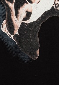 Černé tričko s grafickým potiskem sportovce v akci, podrobně provedené v odstínech hnědé a s texturovaným povrchem.