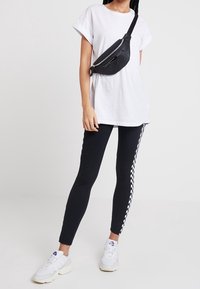 Musta vyölaukku vetoketjuilla, jonka päällä on valkoinen T-paita, yhdistettynä mustiin leggings-housuihin, joissa on ruutukuosi, ja valkoisiin lenkkareihin.