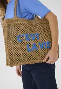 Brun vävd tote bag med blå broderad text "C'EST LA VIE", som visar en öppen vävdesign och hållbara axelremmar.