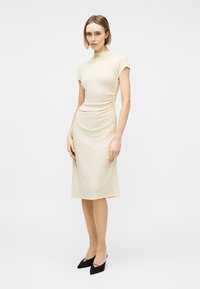 Kvinde iført en beige rynket midi-kjole med kapærmer og høj hals, stylet med sorte spidse hæle, stående på en hvid baggrund.