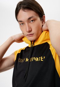 Mustat ja keltaiset urheilulliset hihattomat hupparit, joissa on korkea kaulus ja keltaisella tekstillä "STAY MOTIVATE!". Teksturoitu kangas, jossa on nyöri.