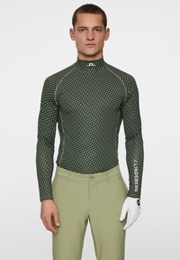 Långärmad golftröja i mörkgrön, med ett geometriskt mönster, hög krage och kontrasterande sömmar. Bärs med ljusgröna byxor och en vit handsk.