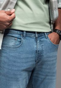 Jeans de mezclilla azul claro con un diseño clásico de cinco bolsillos y una cremallera, con sutiles desvanecimientos y detalles de costura, combinados con una camisa verde.