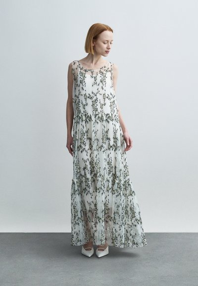 Robe longue blanche à imprimé floral avec des couches translucides, des détails à volants et une silhouette fluide. Associée à des chaussures blanches à bout pointu.