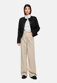 Black quilted cropped jacket with gold buttons, white collared shirt, and beige wide-leg trousers; worn with black shoes.