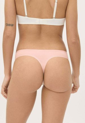 Mujer con sujetador blanco y tanga rosa claro, vista desde atrás, con tatuaje "OHANA" en la parte superior del brazo izquierdo.