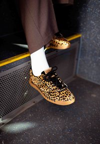 Tenisky se vzorem leopardí kůže s lesklým povrchem, černými tkaničkami a béžovou podrážkou, nošené s hnědými kalhotami a bílými ponožkami, odpočívající na schodu.