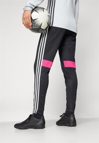 Černé sportovní kalhoty s růžovými akcenty a třemi bílými pruhy po stranách. Na sobě má černé boty, drží černo-bílý fotbalový míč.
