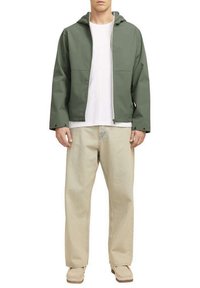 Giacca impermeabile verde con cappuccio, chiusura con zip e bottoni sui lati; indossata sopra una camicia bianca e pantaloni beige larghi. Tessuto morbido.