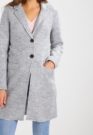 Woman wearing a long gray knit coat with black buttons over a pink top and blue jeans, one hand in coat pocket.