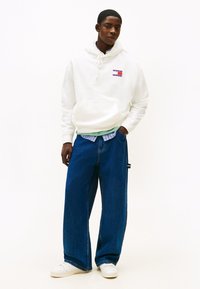 Sudadera con capucha blanca con un bolsillo frontal y un logo, combinada con unos vaqueros azules holgados. Zapatillas blancas completan el conjunto. Fondo blanco liso.