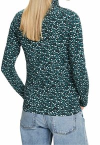 Maglietta a maniche lunghe color teal con un motivo floreale bianco, nero e azzurro chiaro. Presenta un colletto alto e un tessuto morbido e strutturato.