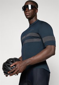 Maillot de cyclisme bleu marine à manches courtes, avec des rayures horizontales grises. Le mannequin tient un casque noir dans une main. Texture de tissu lisse.