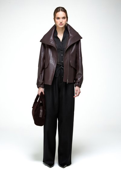 Žena oblečená v príliš veľkej tmavohnedej koženej bunde so širokým golierom, čiernej košeli na gombíky, širokých čiernych nohaviciach a držiaca hnedú chlpatú kabelku.