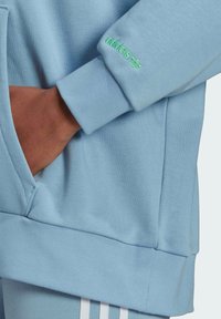 Ljusblå sweatshirt med en ficka, ribbstickade muddar och grön broderad logotyp. Tyget verkar mjukt med en slät struktur.