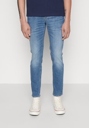 Mann trägt hellblaue Slim-Fit-Jeans und weiße High-Top-Sneaker, steht vor einem schlichten hellen Hintergrund.