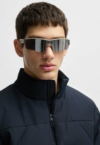 Sunglasses med överdimensionerade grå gradiente linser, en ljusgrå ram och en elegant, aerodynamisk design. Bärs med en svart dragkedjajacka.