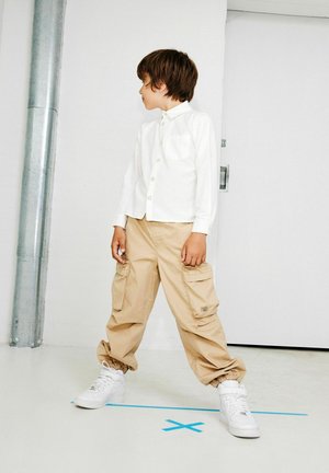 Camicia bianca a maniche lunghe, pantaloni cargo beige con tasche e sneakers bianche. Il bambino si trova su un pavimento chiaro vicino a una parete e un tubo.