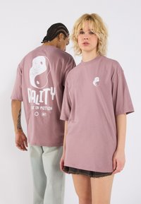 Lilla üle suur t-särk, mille ees ja taga on yin-yang disain, valge tekstiga "KVALITEET" ja "LIIGU LIikumises." Sile puuvilla tekstuur.