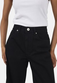 Czarne dżinsowe spodnie o wysokim stanie z bocznymi kieszeniami, zapięcie z przodu na srebrny guzik oraz fakturowana tkanina. Modelka ma na sobie prosty biały top bez rękawów.