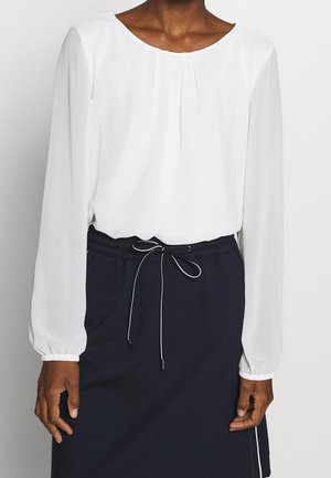 Personne portant un chemisier blanc à manches longues avec des plis, associé à une jupe bleu marine avec une taille à cordon nouée et une bande blanche sur le côté.