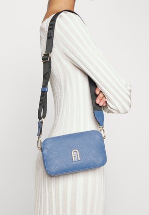 Persoon die een witte geribde jurk draagt en een kleine blauwe leren crossbodytas met een zwarte verstelbare band en zilveren logo aan de voorkant.