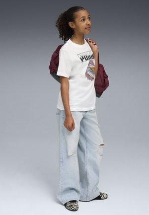 T-shirt blanc avec un design graphique, jean large délavé bleu clair, et chaussures à enfiler rayées noir et blanc. Sac à dos bordeaux sur l'épaule.