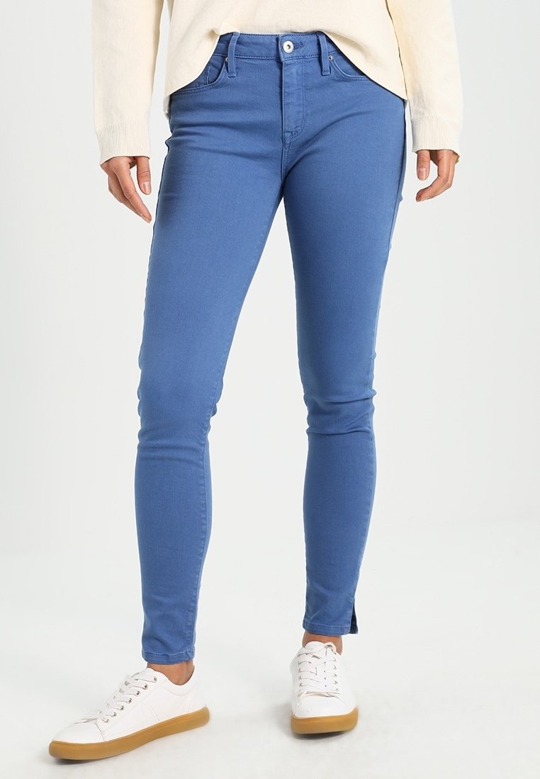 Personne portant un jean slim bleu, un haut blanc à manches longues et des baskets blanches avec des semelles marron, debout sur un fond clair uni.