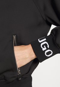 Main dans la poche zippée d'une veste noire, poignet montrant un texte blanc partiel "UGO" sur un tissu côtelé.
