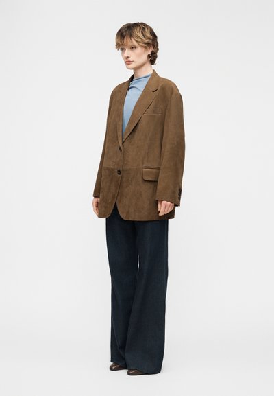 Personne portant un blazer en daim marron surdimensionné, un haut bleu clair, un pantalon large foncé et des chaussures marron, se tenant devant un fond uni.
