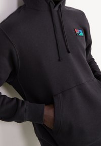 Svart hoodie i mjukt tyg, med en känguruficka, dragsko i huvan och en flerfärgad logotypapplikation på bröstet.