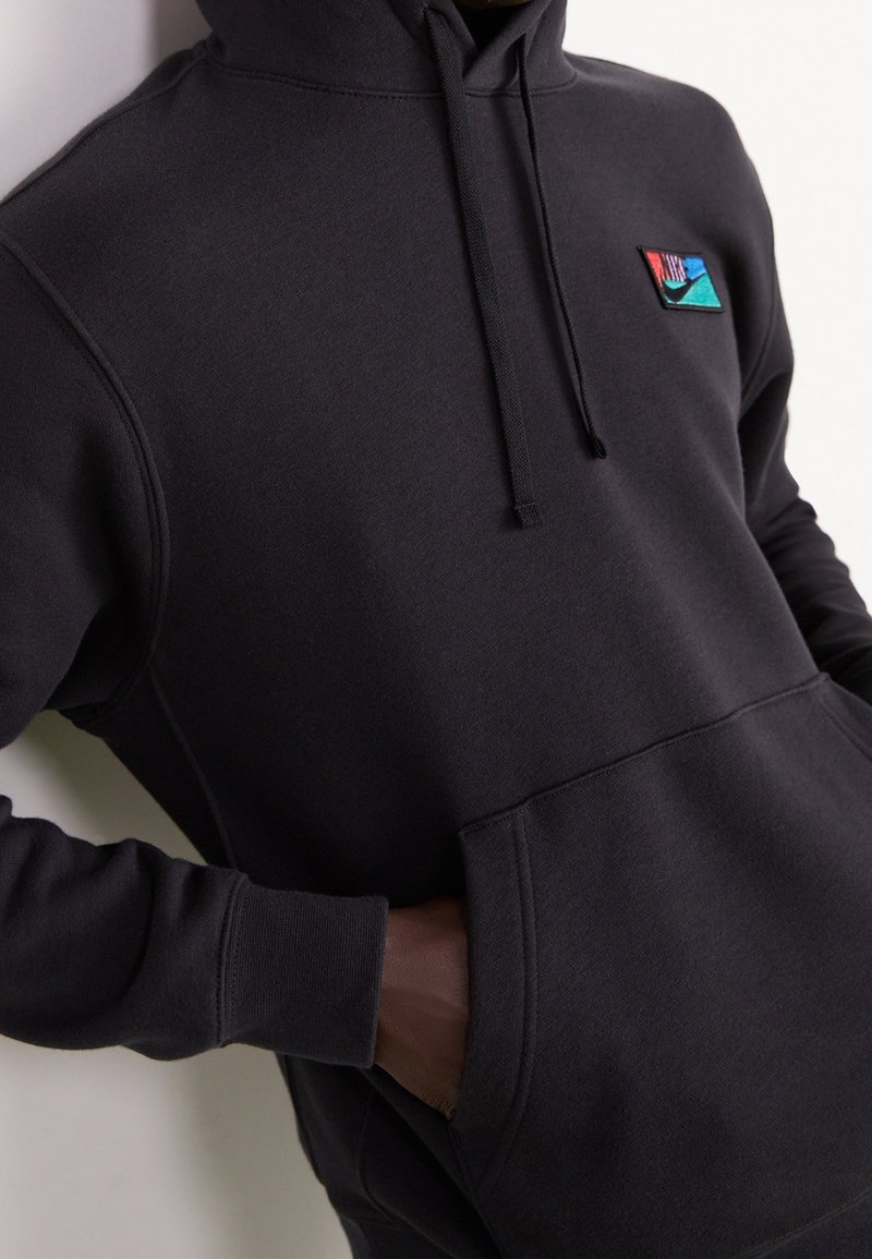 Svart hoodie i mjukt tyg, med en känguruficka, dragsko i huvan och en flerfärgad logotypapplikation på bröstet.