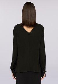 Donna con capelli castani lisci di lunghezza alla spalla, che indossa una blusa nera a maniche lunghe con scollatura a V sul retro, fotografata di spalle.