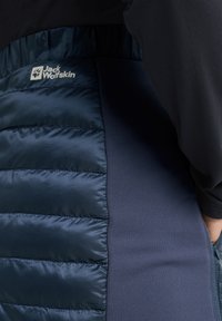 Marine pufferjack met quilted secties aan de ene kant, gladde stof aan de andere kant, en een logo op de bovenste linkerzijde.