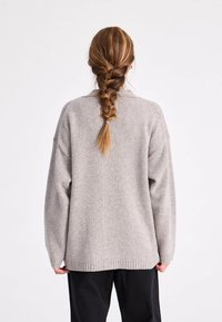 Lichtgrijze gebreide trui met een losse pasvorm, verlaagde schouders en geribbelde zoom. Het model heeft lange gevlochten haren en draagt een zwarte broek.