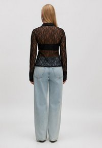 Vrouw met lang blond haar, gekleed in een doorzichtige zwarte kanten top met lange mouwen en hooggenomen lichte blauwe wijde broek, afgebeeld met de rug naar de camera op een effen achtergrond.