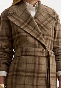 Cappotto di lana a quadri marrone con ampio colletto, vita sblusata e maniche lunghe. Presenta una texture morbida e una camicia bianca a contrasto sotto.