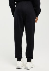 Osoba v černých teplákových kalhotách a bílých teniskách, stojící zády k fotoaparátu před jednoduchým světlým pozadím.