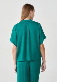 Groene, gestructureerde blouse met een ontspannen pasvorm, korte mouwen en een plooi detail aan de achterkant. De stof lijkt lichtgewicht en vloeiend te zijn.