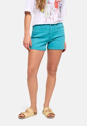 Mujer con pantalones cortos turquesas con dobladillo enrollado, camiseta blanca con gráfico y sandalias beige con hebilla, de pie frente a un fondo liso.