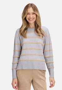 Eine Frau lächelt, trägt einen hellgrauen Pullover mit beigen horizontalen Streifen und beigen Hosen und steht vor einem schlichten weißen Hintergrund.