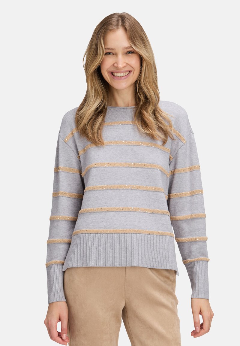 Eine Frau lächelt, trägt einen hellgrauen Pullover mit beigen horizontalen Streifen und beigen Hosen und steht vor einem schlichten weißen Hintergrund.